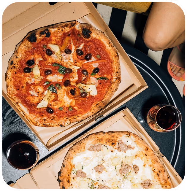 Two artisan pizzas in open boxes on a black table, one veggie one meat, served with red wine